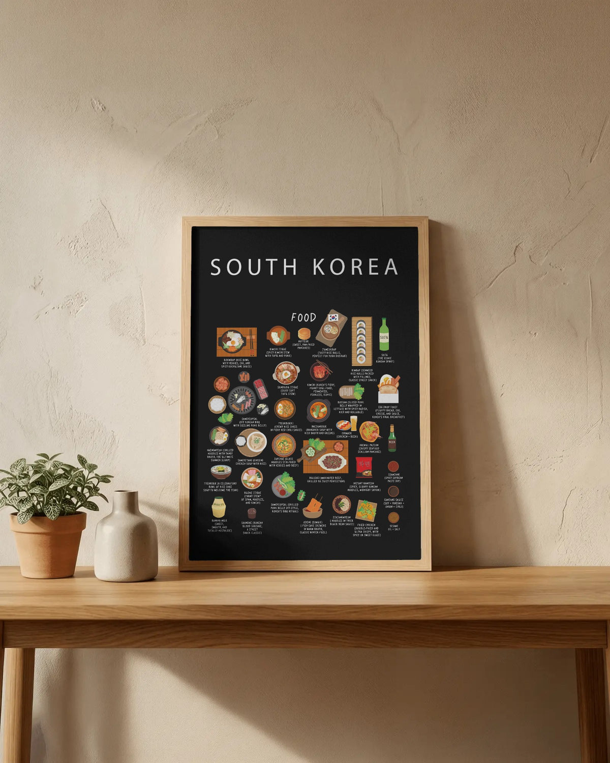 A black South Korea food poster in a light wood frame resting on a wooden table beside a small potted plant and ceramic vase against a textured wall.