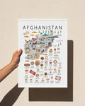 A hand holds a white poster featuring a stylized illustrated map of Afghanistan with food, culture, and landmarks, set against a warm beige wall with a soft shadow.