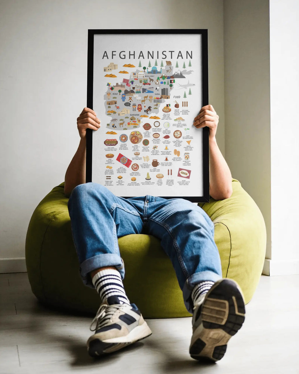 A person sitting in a green bean bag chair holds up a framed travel-style map poster of Afghanistan, with the poster covering their face. The person is wearing blue jeans, striped socks, and sneakers in a minimalist indoor room.