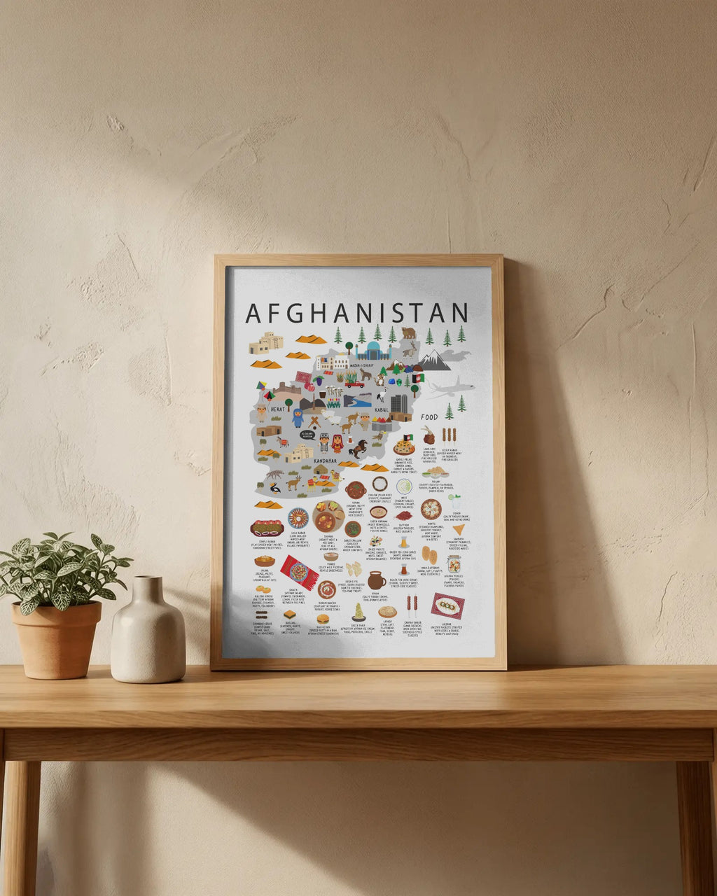A framed travel-style map poster of Afghanistan with many illustrated landmarks and objects, leaning on a wooden table against a textured beige wall, with small potted plants beside it.