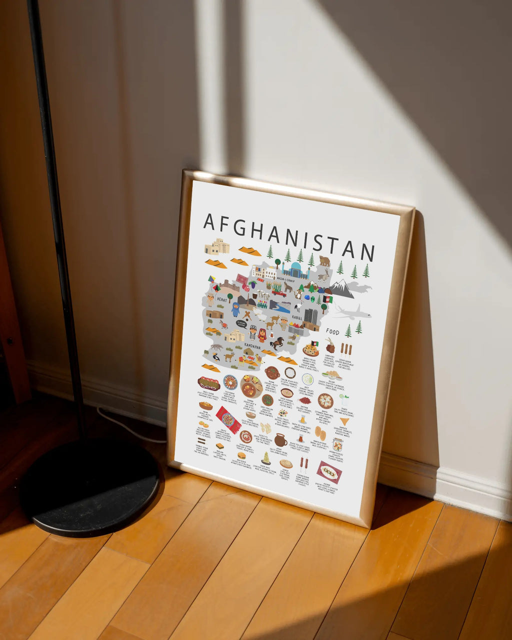 A framed travel-style map poster of Afghanistan leaning against a wall on a wooden floor, with strong sunlight casting shadows across the room and a black floor lamp visible at the left edge.