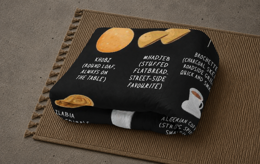 A folded black blanket or throw featuring a printed food and drink theme with white text labels and illustrations of flatbread, a croquette, and a coffee cup, placed on a woven brown mat over a gray floor.