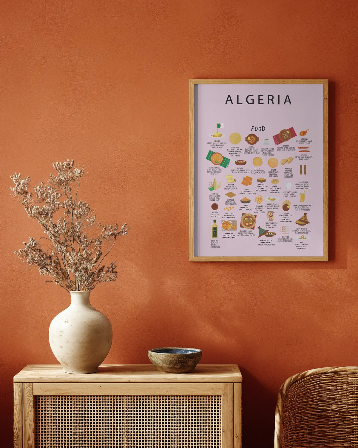 A framed poster titled 'ALGERIA FOOD' hanging on a terracotta-colored wall above a wooden cabinet with a vase of dried flowers and a small bowl.