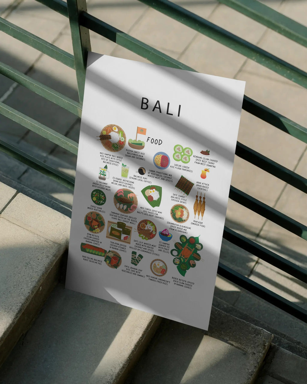 A white Bali food poster resting on outdoor concrete steps against a green metal railing, cast in soft sunlight and shadows.