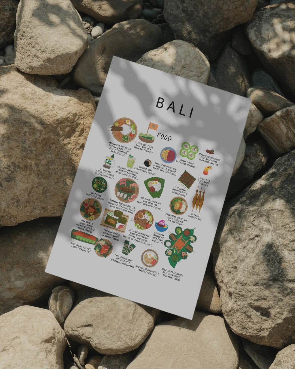 An unframed white poster featuring Balinese food illustrations laid out on a bed of natural smooth rocks in dappled sunlight.