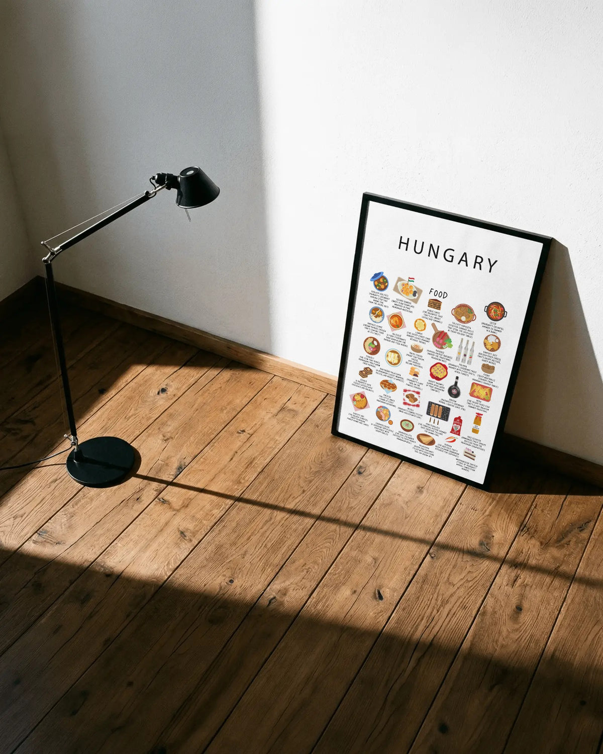 A framed white Hungary food poster leans against a white wall on a natural wood floor, beside a black adjustable floor lamp illuminated by sunlight.