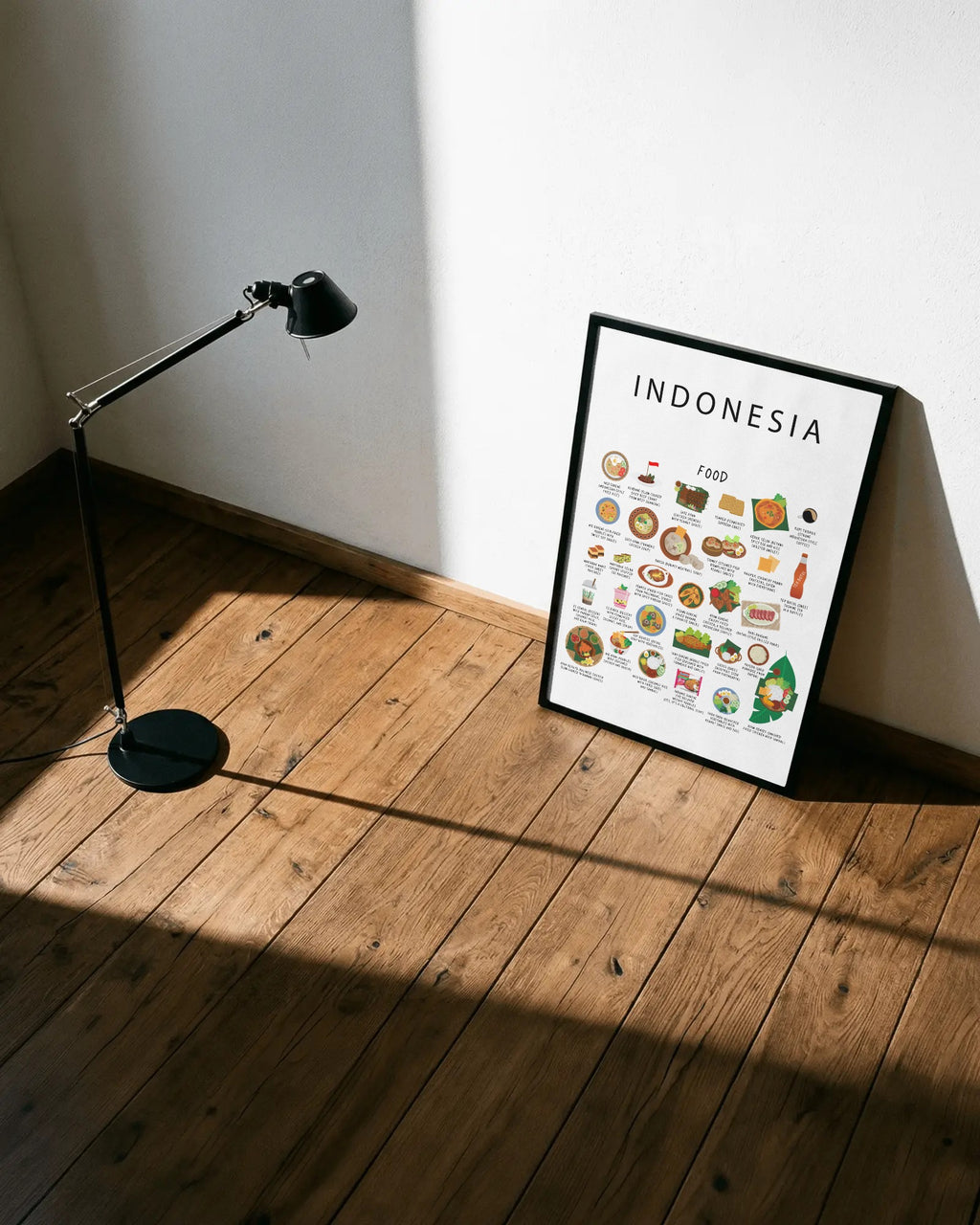 A framed white Indonesian food poster rests on wooden floorboards against a white wall, positioned beside a black adjustable floor lamp.