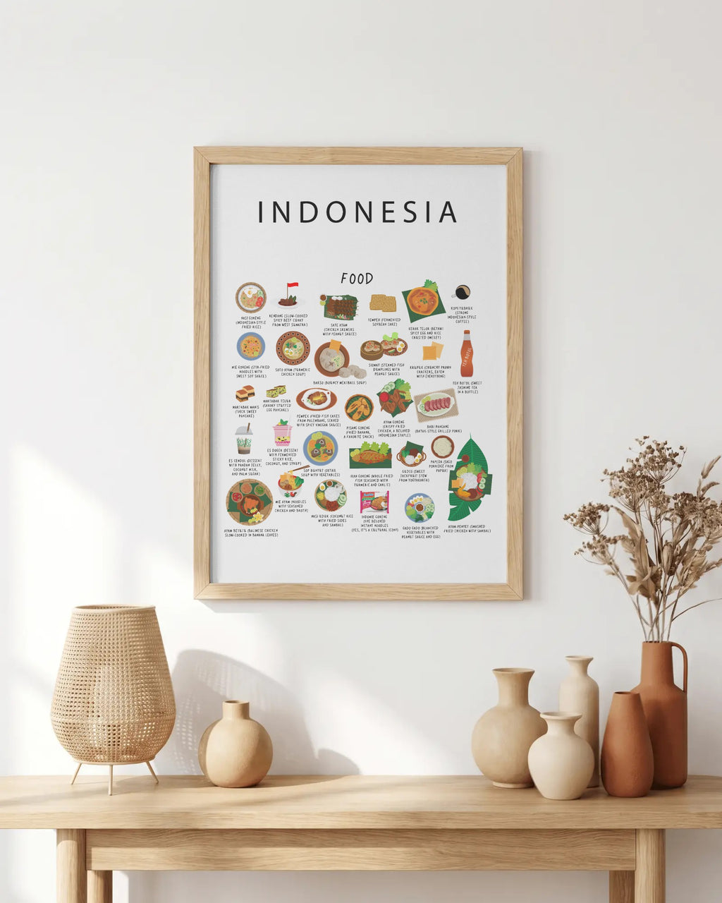 An Indonesian food poster in a light wood frame hangs on a white wall above a wooden table with ceramic vases and dried florals.