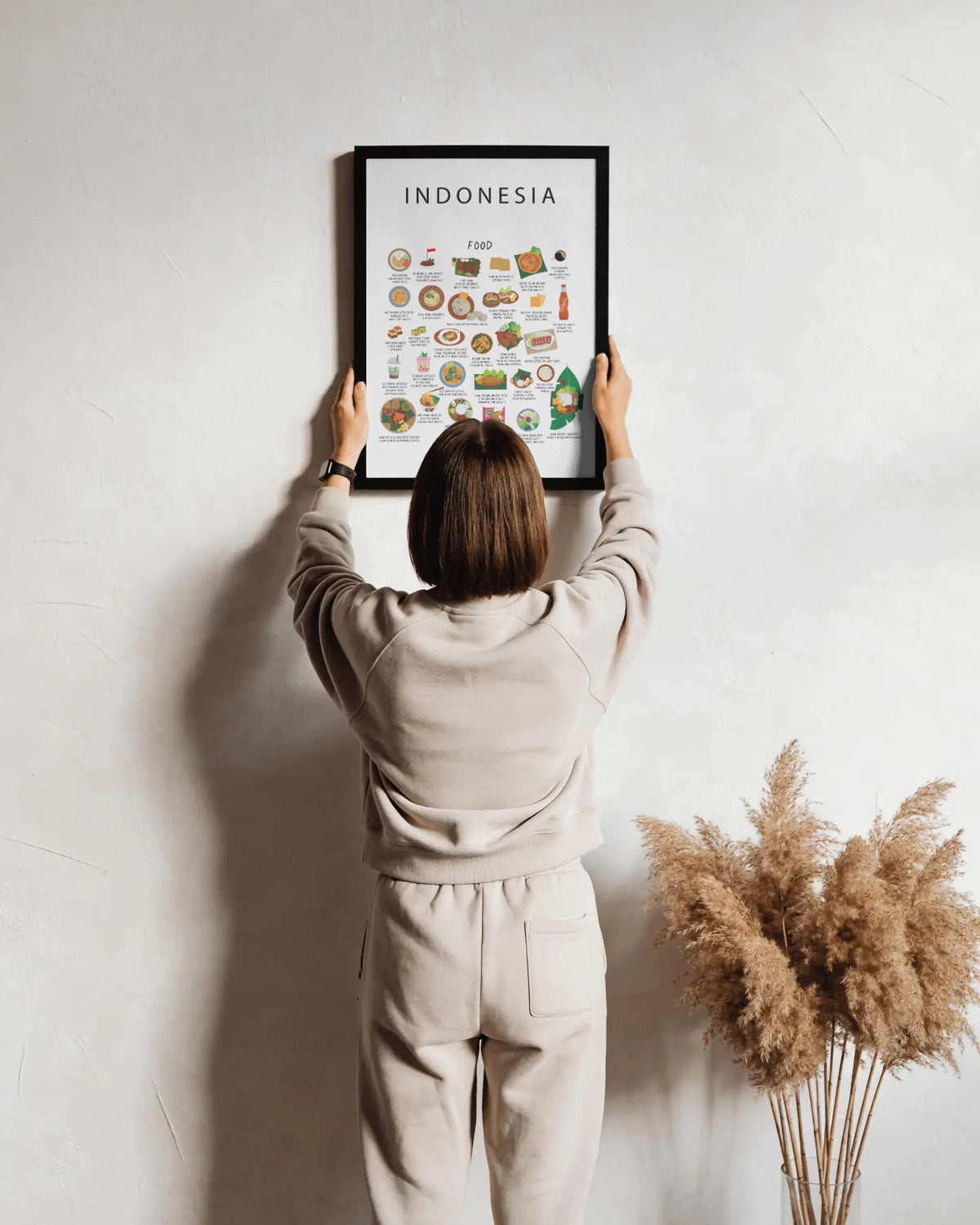 A person hangs a black-framed white Indonesia food poster on a textured wall beside a vase of dried grass.