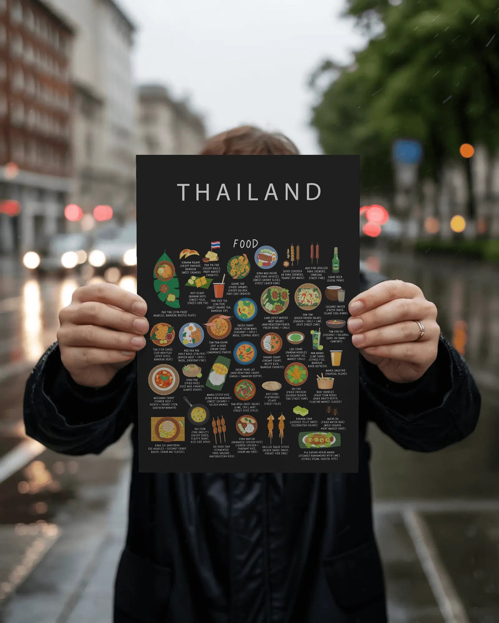A person holds a black Thailand food poster in front of their face against a blurred city street with glowing vehicle lights.