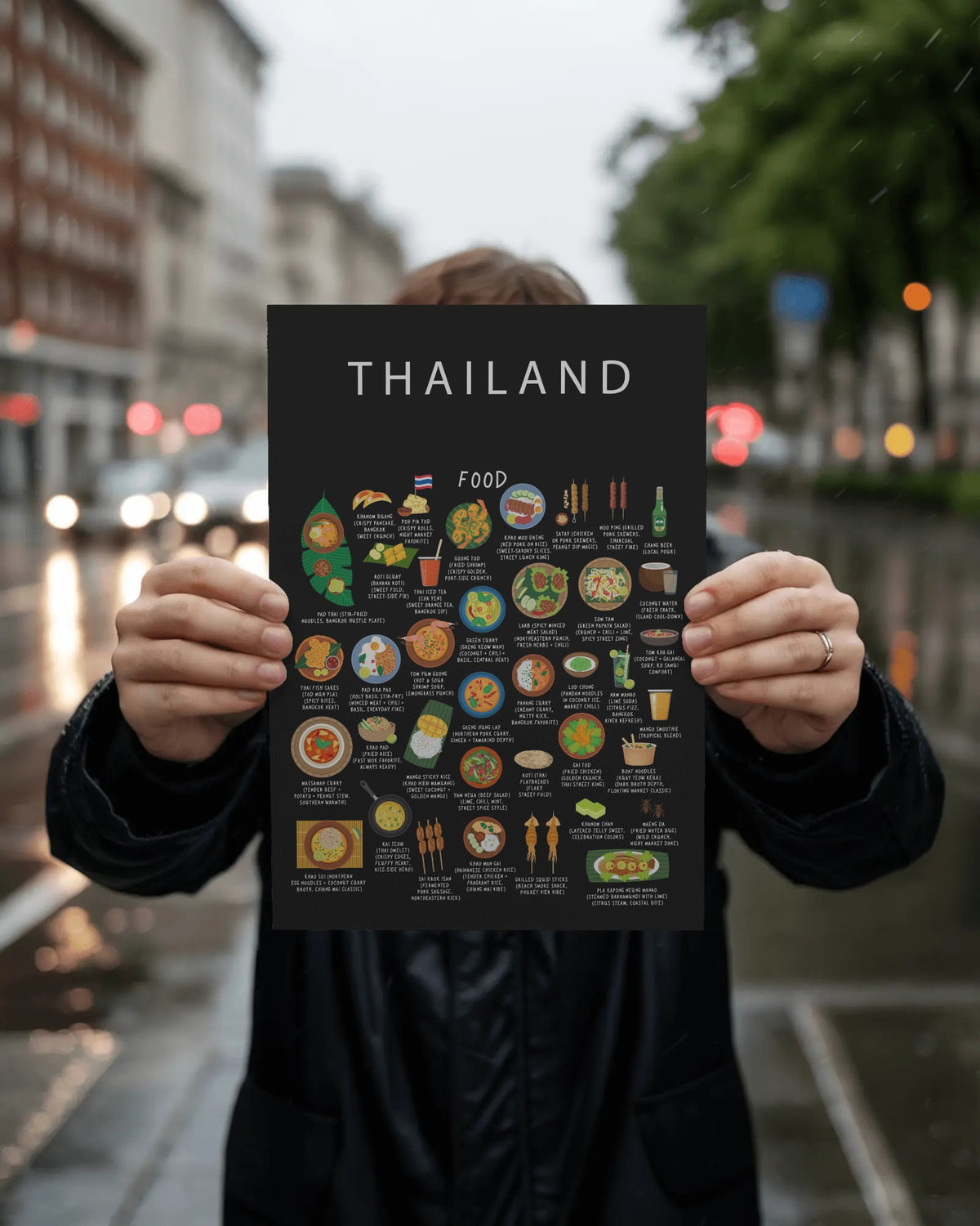 A person holds a black Thailand food poster in front of their face against a blurred city street with glowing vehicle lights.