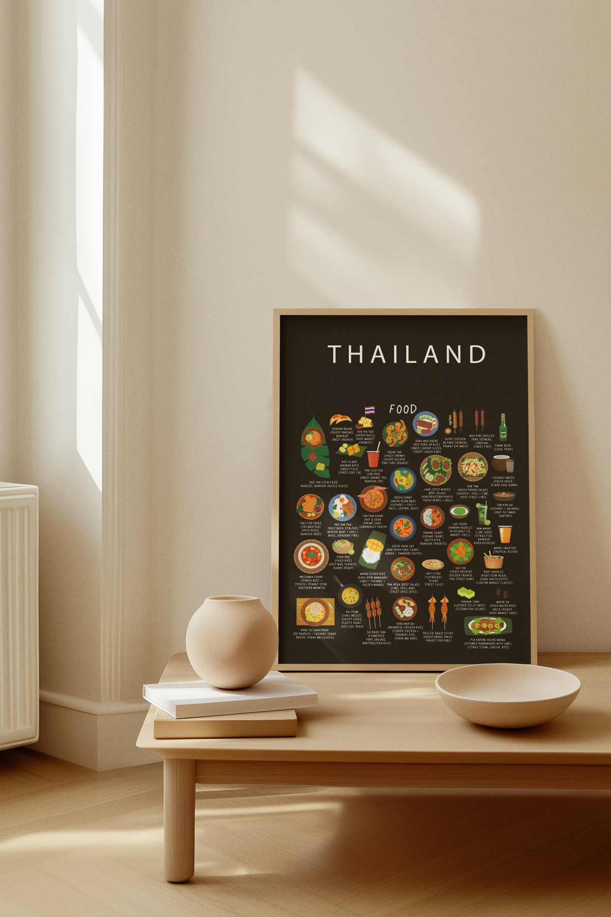 A black Thailand food poster in a light wood frame rests on a wooden bench beside a round ceramic vase on books and a shallow bowl.