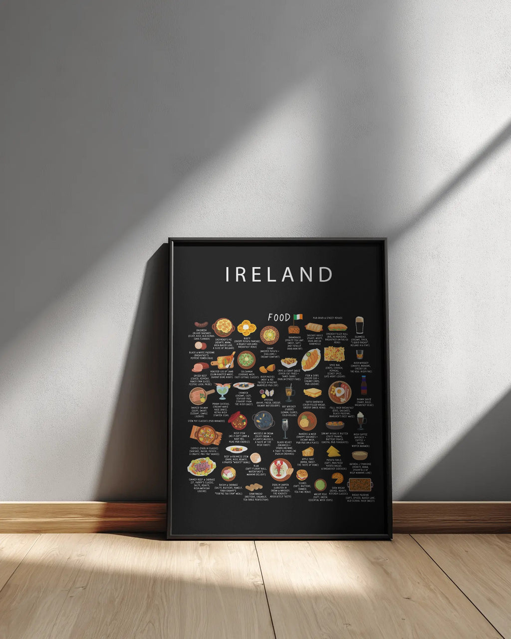 A framed black Ireland food poster rests on a light wood floor, leaning against a neutral grey wall illuminated by diagonal sunlight and shadows.