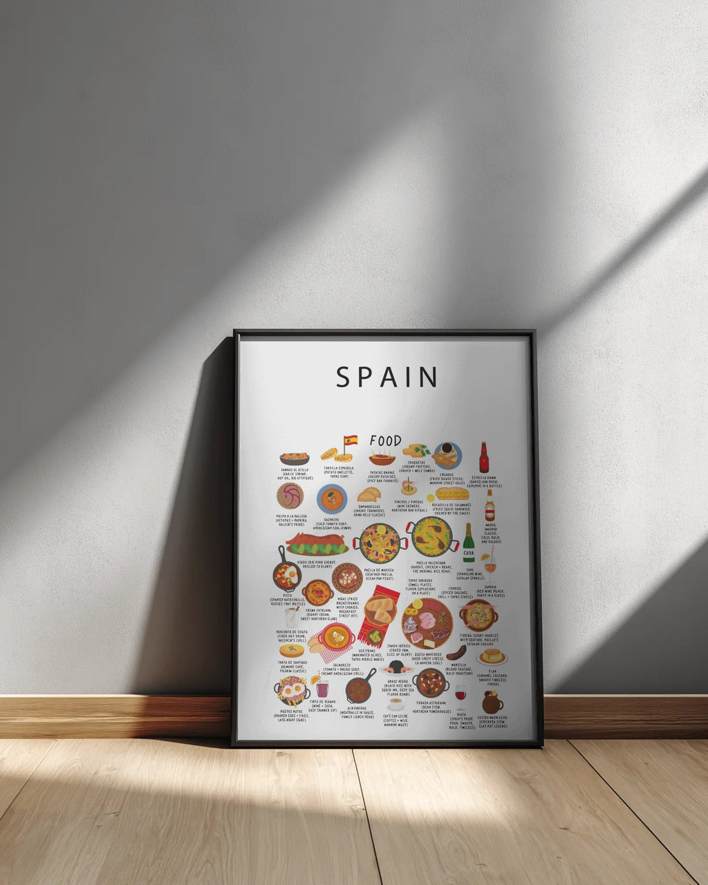 A black-framed Spain food poster with illustrated dishes rests on a light wooden floor against a neutral wall with diagonal sunlight shadows.