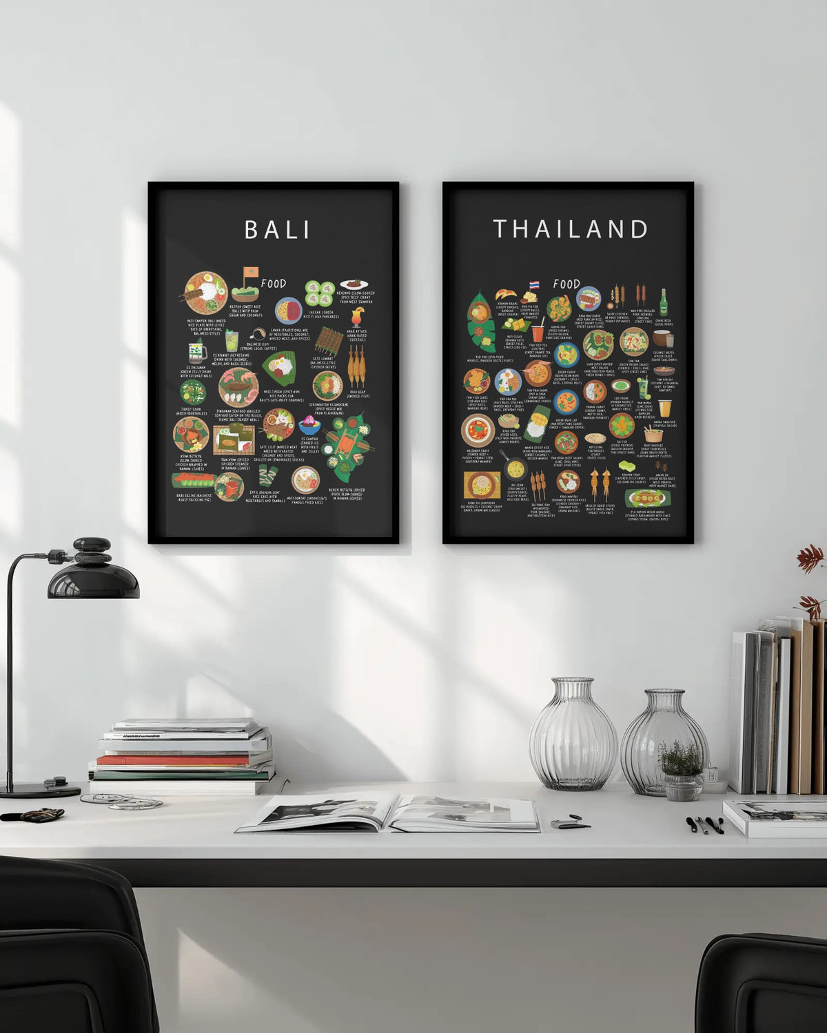 A framed black Thailand food poster hanging beside a Bali poster on a white wall above a modern desk with a lamp, books, and glass vases.
