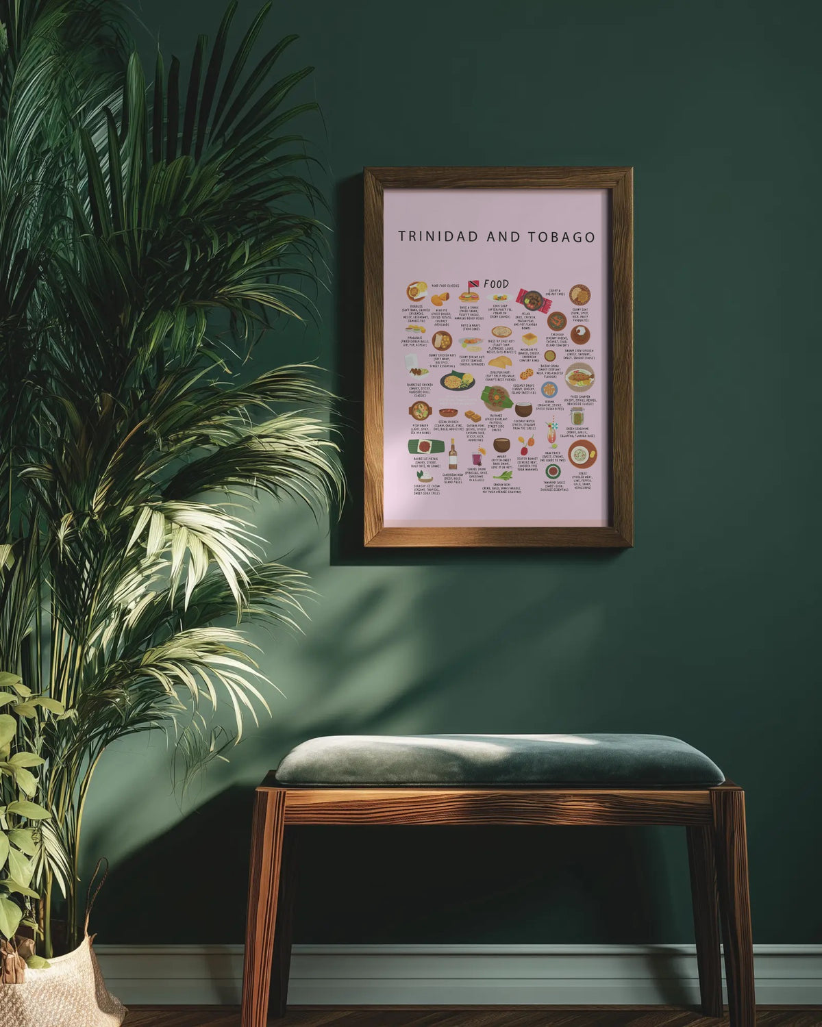 Framed pink Trinidad food poster hanging on a dark green wall above a wooden bench with a cushioned seat, next to a large potted palm plant.